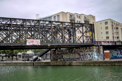 Frédéric · 13/09/2025 · 2518 x 1680 Cette photographie montre un **pont métallique** enjambant un canal, avec des **bâtiments urbains** en arrière-plan. Le sujet principal est le pont, marqué par des **graffitis** et une signalétique rouge ("Attention, bas niveau d'eau").La **composition** est équilibrée, avec une perspective légèrement basse. Le **cadrage** inclut une partie de l’eau et des bâtiments, créant une ambiance urbaine et industrielle. La **lumière** est naturelle mais tamisée, donnant une teinte grise et froide. Les **couleurs dominantes** sont les tons gris, verts (eau) et ocres (bâtiments), avec des touches de rouge et de noir (graffitis).Le style est **réaliste et documentaire**, évoquant une ambiance **urbaine et légèrement négligée**.