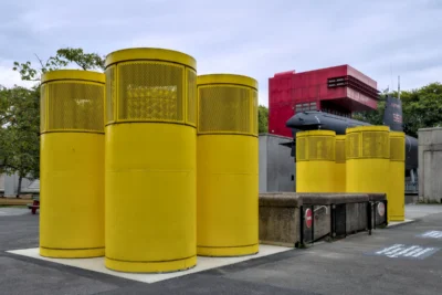 Frédéric · 13/09/2025 · 2517 x 1680 Cette photographie montre trois grands cylindres jaunes empilés, probablement des poubelles industrielles, placées sur un parking. En arrière-plan, on aperçoit des conteneurs métalliques et une structure rouge.La composition est épurée, avec un cadrage serré sur les cylindres. La lumière est naturelle, diffuse, sans ombres marquées, suggérant un jour nuageux. Les couleurs dominantes sont le jaune vif des cylindres et le gris des surfaces environnantes, avec une touche de rouge en arrière-plan.Le style est documentaire, réaliste, et l’ambiance industrielle et utilitaire.