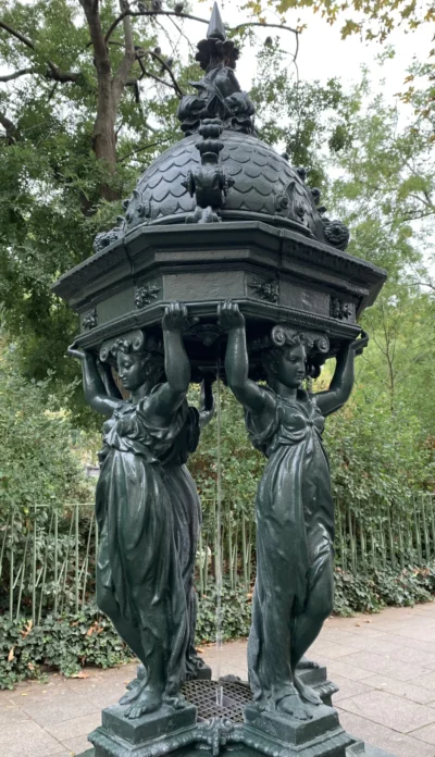 Kerry · 13/09/2025 · 2270 x 3947 Cette photographie montre une fontaine en bronze représentant un groupe de figures féminines soutenant une vasque ornée. Le sujet principal est ce monument sculptural, richement détaillé, avec des motifs végétaux et des personnages élégants.La composition est symétrique et verticale, mettant en valeur la structure et les détails des sculptures. Le cadrage est serré, centré sur la fontaine, avec un arrière-plan flou de végétation. La lumière naturelle, douce et diffuse, accentue les contours et les textures du bronze. Les couleurs dominantes sont les verts des feuilles et le gris-bleuté du bronze, créant une ambiance à la fois classique et paisible.