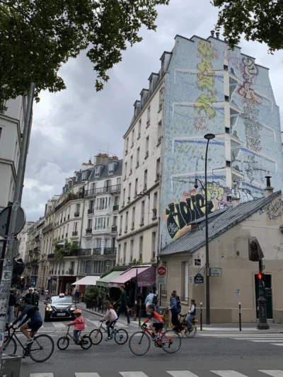 Kerry · 13/09/2025 · 2393 x 3190 Cette photographie capture une rue parisienne animée. Le **sujet principal** est un immeuble aux **fresques murales colorées**, représentant des personnages stylisés. La **composition** montre une perspective en contre-plongée, avec des cyclistes et des piétons en mouvement devant.Le **cadrage** est serré sur l’immeuble, encadré par des bâtiments classiques. La **lumière** est naturelle, légèrement voilée par un ciel nuageux, créant une ambiance douce. Les **couleurs dominantes** sont les tons pastel des fresques, contrastant avec les gris des bâtiments et le vert des arbres.Le style est **urbain et artistique**, évoquant une ambiance **vivante et créative**, typique des quartiers dynamiques de Paris.