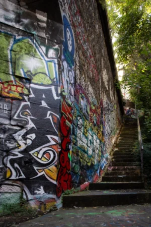 Claude · 2632 x 3948 Cette photographie met en scène un **mur tagué** couvert de graffitis colorés et variés, encadrant un escalier en pierre. Le **sujet principal** est l’art urbain, avec des motifs stylisés et des couleurs vives.La **composition** joue sur la perspective, avec un **cadrage serré** qui guide le regard vers les marches. La **lumière**, naturelle et tamisée, crée des contrastes entre ombres et éclats colorés. Les **couleurs dominantes** sont vives (rouge, bleu, jaune) mais contrastées par des teintes sombres.Le **style** est urbain et spontané, évoquant une **ambiance** à la fois dynamique et mystérieuse, typique des espaces street art.