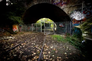 Claude · 5824 x 3883 Cette photographie montre un tunnel ferroviaire abandonné, sujet principal. La composition met en valeur les rails centraux, encadrés par des graffitis colorés sur les murs et une clôture métallique.Le cadrage est serré, guidant le regard vers l’obscurité au loin. La lumière, tamisée et contrastée, crée une ambiance mystérieuse et dramatique. Les couleurs dominantes sont les tons sombres (noir, gris) rehaussés par des éclats de couleurs vives des graffitis (rouge, bleu, jaune).Le style est réaliste et légèrement cinématographique, évoquant une atmosphère abandonnée et mélancolique.