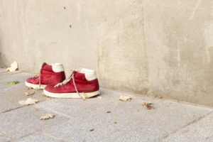Claude · 4059 x 2706 Cette photographie met en scène une paire de **sneakers rouges** avec des semelles blanches, posées sur un **sol en pierre**. Le sujet principal est ces chaussures, légèrement usées, avec des lacets défaits.La **composition** est épurée, centrée sur les chaussures, avec un arrière-plan neutre en **mur beige**. Le **cadrage** est serré, mettant en valeur les détails des chaussures et des feuilles mortes au sol.La **lumière** semble naturelle, douce et légèrement tamisée, créant des ombres subtiles. Les **couleurs dominantes** sont le rouge vif des chaussures et le beige du mur, renforçant un style **minimaliste et urbain**. L’ambiance est **calme et mélancolique**, évoquant l’abandon ou la nostalgie.