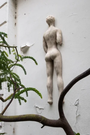 Claude · 3375 x 5063 Cette photographie met en scène une sculpture en relief représentant une silhouette féminine stylisée, apposée sur un mur blanc délavé. Le cadrage serré isole la figure, encadrée par une branche d'arbuste en bas à gauche.La lumière naturelle, douce et légèrement tamisée, accentue les contours et les textures. Les couleurs dominantes sont les tons pastel (blanc, beige) et les verts clairs de la plante. Le style est épuré et minimaliste, évoquant une ambiance à la fois moderne et intemporelle, presque poétique et mélancolique.