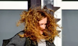 Franck T. · 04/10/2025 · 1000 x 588 Cette photographie met en scène une femme au premier plan, dont les cheveux blonds et bouclés sont soulevés par le vent. Le cadrage est serré sur son visage et ses cheveux, créant un effet dynamique.La composition est simple, avec un arrière-plan flou et géométrique, probablement une structure architecturale. La lumière naturelle semble venir de la gauche, éclairant le sujet de manière contrastée. Les couleurs dominantes sont les tons chauds (blond, beige) et les gris froids de l’arrière-plan.Le style est naturel et spontané, évoquant une ambiance vivante et énergique.