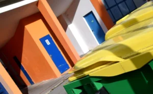 Franck T. · 04/10/2025 · 1000 x 619 Cette photographie met en scène un bâtiment aux couleurs vives avec des portes orange et bleue. Le sujet principal semble être les bannières ou enseignes jaunes et vertes en avant-plan, partiellement floues, créant un effet dynamique.La composition joue sur les contrastes entre les angles géométriques du bâtiment et les formes arrondies des enseignes. Le cadrage est serré, avec une perspective légèrement inclinée. Les couleurs dominantes sont le jaune, l’orange, le bleu et le vert, apportant une ambiance moderne et énergique. La lumière semble naturelle, mettant en valeur les teintes vives et les ombres douces.