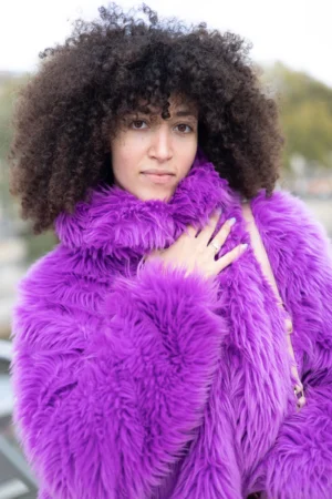 Claude · 4000 x 6000 Cette photographie met en scène une femme comme sujet principal, avec une coiffure afro volumineuse et un large manteau en fourrure synthétique violet. Le cadrage est serré, centré sur son visage et sa tenue, avec un arrière-plan flou et légèrement délavé.La lumière semble naturelle, douce et légèrement tamisée, créant des ombres subtiles. Les couleurs dominantes sont le violet éclatant du manteau et les tons neutres de sa peau et de l’arrière-plan. Le style est à la fois streetwear et avant-gardiste, évoquant une ambiance moderne et audacieuse.