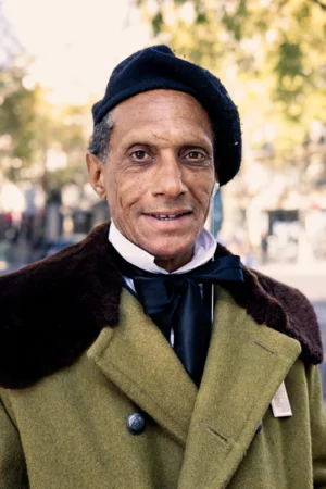 Claude · 4000 x 6000 Cette photographie met en scène un homme âgé comme sujet principal, vêtu d’un costume traditionnel avec un long manteau vert et un chapeau noir. Le cadrage est serré sur son visage et sa poitrine, isolant le sujet de l’arrière-plan flou.La lumière semble naturelle, douce et légèrement tamisée, créant des ombres subtiles. Les couleurs dominantes sont les tons verts du manteau, les gris et noirs du costume, ainsi que les teintes chaudes de la peau et du col blanc.Le style évoque une touche documentaire ou historique, avec une ambiance sobre et chaleureuse, renforcée par les feuilles automnales en arrière-plan.