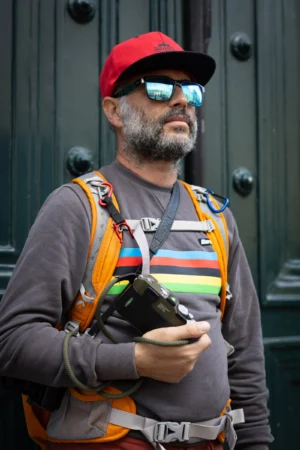 Claude · 3389 x 5083 Cette photographie montre un homme comme sujet principal, vêtu d’un gilet de sauvetage coloré et d’un bonnet rouge, tenant un appareil photo. La composition est centrée sur son visage et son torse, avec un cadrage serré.La lumière semble naturelle, légèrement contrastée. Les couleurs dominantes sont les tons vifs du gilet (orange, jaune, vert) et le gris du vêtement. Le style est documentaire et spontané, capturant une scène réaliste. L’ambiance est à la fois sérieuse et décontractée, évoquant une activité professionnelle ou un loisir lié à la photographie.