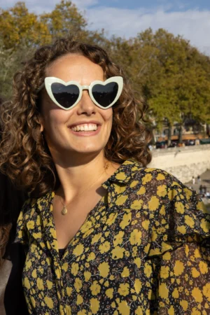Claude · 3286 x 4929 Cette photographie met en scène une femme souriante, sujet principal, avec des lunettes de soleil à monture blanche. Elle porte une chemise à motifs floraux jaunes et noirs. Le cadrage est serré, centré sur son visage et sa poitrine.La lumière semble naturelle et douce, probablement en extérieur. Les couleurs dominantes sont les tons chauds (jaune, vert des arbres en arrière-plan) et les contrastes doux.Le style est décontracté et estival, évoquant une ambiance détendue et joyeuse, probablement prise lors d’une journée ensoleillée en plein air.