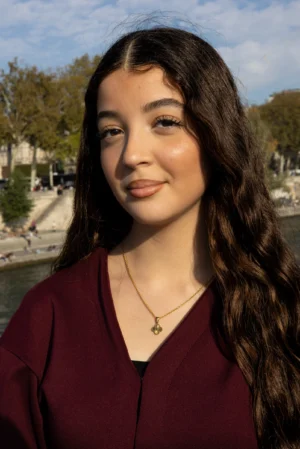 Claude · 3747 x 5613 Cette photographie met en scène une jeune femme comme sujet principal. Le cadrage est serré sur son visage et sa poitrine, avec un arrière-plan flou montrant un paysage urbain et des arbres.La lumière est naturelle et douce, éclairant son visage de manière homogène. Les couleurs dominantes sont les tons chauds de sa tenue marron-rouge et ses cheveux foncés, contrastant avec le bleu du ciel.Le style est simple et naturel, évoquant une ambiance détendue et authentique.