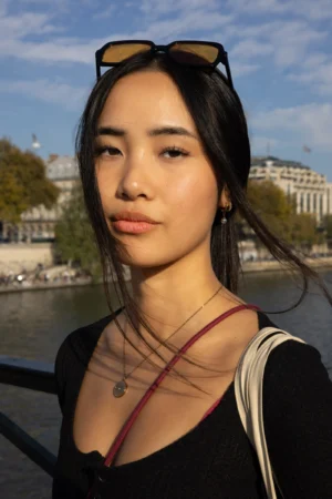 Claude · 3911 x 5866 Cette photographie met en scène une jeune femme comme sujet principal, posée devant un cadre urbain flou. Le cadrage est serré sur son visage et sa poitrine, avec un fond de rivière et de bâtiments.La lumière est naturelle, douce et légèrement latérale, créant des ombres subtiles. Les couleurs dominantes sont les tons chauds (peau, rouge du collier) contrastant avec les teintes froides du fond (bleu du ciel et de l’eau).Le style est épuré et contemporain, évoquant une ambiance à la fois calme et légèrement mystérieuse.