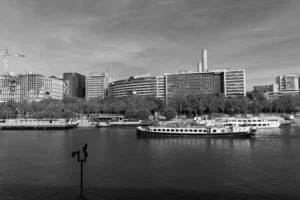 Claude · 5639 x 3759 Cette photographie en noir et blanc met en scène un **port urbain** avec un **bateau à passagers** comme sujet principal. La composition montre une **perspective large**, incluant des bâtiments modernes en arrière-plan et des arbres le long des quais.Le **cadrage** est équilibré, avec une ligne d’horizon centrale. La lumière, douce et diffuse, crée des contrastes modérés, accentuant les formes architecturales. Les **couleurs dominantes** sont les nuances de gris, renforçant l’atmosphère classique et intemporelle.Le style est **documentaire et esthétique**, évoquant une ambiance **calme et urbaine**, typique des villes portuaires.