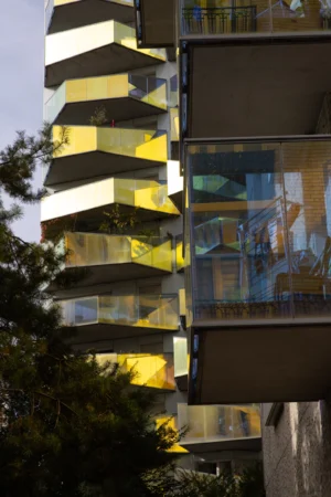 Claude · 3813 x 5719 Cette photographie met en scène un immeuble moderne aux formes géométriques anguleuses. Le sujet principal est la façade architecturale, composée de balcons en saillie et de grandes surfaces vitrées. Le cadrage est serré, mettant en valeur les jeux de lumière et d’ombre.Les couleurs dominantes sont les tons gris, jaunes et dorés, accentués par les reflets du soleil. La lumière naturelle crée des contrastes marqués et des éclats lumineux. Le style est contemporain et minimaliste, évoquant une ambiance futuriste et urbaine. Les arbres en bas de l’image apportent une touche naturelle contrastant avec l’architecture.