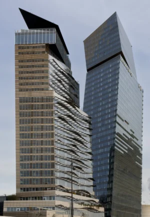 Claude · 3231 x 4662 Cette photographie met en scène deux gratte-ciels modernes comme sujets principaux. La composition est symétrique, avec les bâtiments se faisant face, créant une impression de dualité. Le cadrage est serré, mettant en valeur leurs formes architecturales distinctes et leurs façades en verre et métal.La lumière, douce et naturelle, semble provenir d’un ciel légèrement nuageux, adoucissant les contours. Les couleurs dominantes sont les tons gris et bleus métallisés, reflétant une ambiance urbaine et contemporaine. Le style architectural est résolument moderne et minimaliste, évoquant puissance et innovation. L’ambiance est à la fois dynamique et froide, typique des centres urbains modernes.