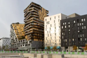 Claude · 4000 x 2667 Cette photographie montre un ensemble d'immeubles modernes en milieu urbain. Le sujet principal est un groupe de bâtiments aux architectures contemporaines, avec des façades variées : bois, béton et verre.La composition met en valeur des lignes verticales et horizontales, créant un jeu de volumes. Le cadrage est serré, centré sur les structures. La lumière, naturelle et diffuse, adoucit les contrastes. Les couleurs dominantes sont les tons neutres (gris, beige) rehaussés par des touches de bois chaud et de noir.Le style est épuré et architectural, évoquant une ambiance urbaine dynamique et moderne.