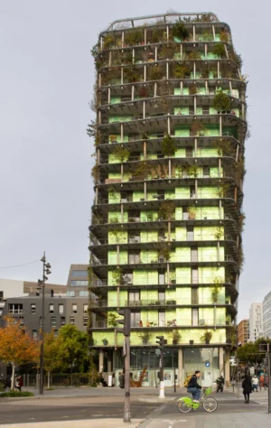 Claude · 3331 x 5264 Cette photographie met en scène un immeuble moderne recouvert de végétation, sujet principal. La composition montre un cadrage serré sur le bâtiment, avec des arbres et des passants en bas, créant un contraste entre nature et architecture urbaine. La lumière est naturelle, légèrement diffuse, mettant en valeur les détails de la façade.Les couleurs dominantes sont les verts de la végétation, les tons gris de la structure et des éléments urbains, avec des touches de blanc et de bleu clair. Le style est contemporain et innovant, évoquant une ambiance futuriste et écologique.