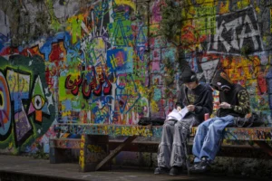 Thierry · 04/10/2025 · 3840 x 2560 Cette photographie met en scène un homme assis sur un banc, absorbé par un livre ou un appareil photo. Le sujet principal est donc cet individu, dont le visage est partiellement caché par une casquette.La composition joue sur le contraste entre le calme du personnage et le mur coloré de graffitis derrière lui. Le cadrage est serré, centré sur le banc et l’homme, avec une profondeur de champ réduite.Les couleurs dominantes sont vives et variées grâce aux graffitis, tandis que l’homme est habillé de tons neutres. La lumière semble naturelle, tamisée, créant une ambiance à la fois urbaine et paisible. Le style est réaliste et documentaire.