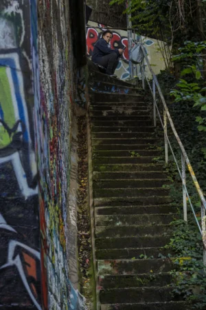 Thierry · 04/10/2025 · 2560 x 3840 Cette photographie met en scène un escalier en pierre couvert de mousse, encadré par un mur tagué. Le sujet principal est l’escalier, qui mène vers une lumière tamisée en haut à gauche.La composition est asymétrique, avec un cadrage serré sur les marches et une partie du graffiti coloré. La lumière, douce et naturelle, semble filtrer à travers les feuilles, créant des ombres douces. Les couleurs dominantes sont les teintes vives des tags (bleu, rouge, vert) contrastant avec les tons verts de la végétation et les gris de la pierre.L’ambiance est à la fois urbaine et naturelle, mélancolique et artistique.