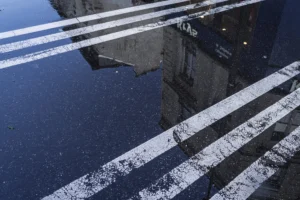 Thierry · 04/10/2025 · 3840 x 2560 Cette photographie montre principalement une portion de trottoir et de chaussée avec des marquages blancs usés et une flaque d’eau reflétant un pont ou une structure en béton. La composition repose sur une perspective basse, accentuant les lignes horizontales et verticales. Le cadrage est serré, centré sur les détails des marquages et de la réflexion.Les couleurs dominantes sont les tons gris et bleu, avec des reflets sombres. La lumière semble naturelle, probablement en journée, créant des contrastes nets. Le style est épuré et réaliste, évoquant une ambiance urbaine et légèrement mélancolique, soulignée par la monotonie des éléments architecturaux.