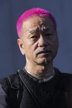 Thierry · 07/10/2025 · 2560 x 3840 Cette photographie met en scène un homme au regard intense, sujet principal. Son visage est partiellement éclairé, avec une lumière contrastée qui accentue ses traits. Il arbore une coiffure teinte en violet et une barbe grisonnante.Le cadrage est serré, centré sur son visage, créant une ambiance intime. Les couleurs dominantes sont les tons froids du fond et les teintes chaudes de sa peau et de ses cheveux violets. Le style semble naturel mais expressif, évoquant une ambiance à la fois mystérieuse et puissante.