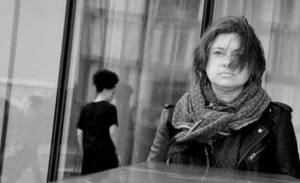 Franck T. · 18/10/2025 · 1000 x 611 Cette photographie en noir et blanc met en scène une femme comme sujet principal, vêtue d’un manteau et d’un long écharpe, regardant pensivement à travers une vitre. La composition joue sur le contraste entre elle et son reflet flou à l’intérieur. Le cadrage serré isole son visage, renforçant l’introspection.La lumière douce et tamisée crée une ambiance mélancolique et contemplative. Les tons dominants, bien que monochromes, varient du clair au foncé, accentuant les textures et les émotions. Le style sobre et réaliste, typique du photojournalisme, renforce une atmosphère à la fois intime et universelle.