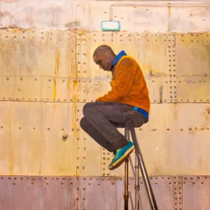 Jean-Paul · 16/10/2025 · 1956 x 1956 Cette photographie met en scène un homme assis sur un tabouret métallique, adossé à une paroi industrielle. Le sujet principal est le visage de l’homme, en contre-plongée, ce qui accentue son expression mélancolique.La composition est épurée, avec un cadrage serré. La lumière naturelle, douce et légèrement rasante, crée des ombres marquées. Les couleurs dominantes sont des tons neutres (beige, gris) avec des touches de bleu et orange (vêtements).Le style est réaliste et documentaire, évoquant une ambiance contemplative et presque urbaine, teintée de solitude.