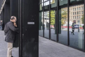 Benoit · 18/10/2025 · 6932 x 4621 Cette photographie montre un homme, sujet principal, discutant au téléphone près d’une entrée vitrée. La composition met en avant un cadrage serré sur lui, avec des reflets dans les vitres et des bâtiments modernes en arrière-plan.La lumière est naturelle, probablement en journée, avec des ombres douces. Les couleurs dominantes sont les tons gris (bâtiments, vêtements) et quelques touches de vert (végétation) et rouge (signalisation).Le style est réaliste et documentaire, capturant une scène urbaine banale. L’ambiance est calme et contemporaine, reflétant le quotidien en ville.