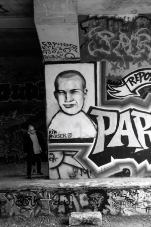 Philippe · 04/10/2025 · 2001 x 3000 Cette photographie en noir et blanc met en scène un graffiti comme sujet principal, représentant un personnage avec le mot "PAPA" stylisé. La composition inclut une personne en contrebas, à gauche, ajoutant une échelle humaine.Le cadrage est serré sur le graffiti, avec des éléments architecturaux en arrière-plan. La lumière est naturelle, contrastée, accentuant les textures et les détails. Les couleurs dominantes sont les nuances de gris, renforçant l’aspect urbain et street art. Le style est brut et expressif, typique des tags. L’ambiance est à la fois sombre et dynamique, mêlant street culture et intimité.