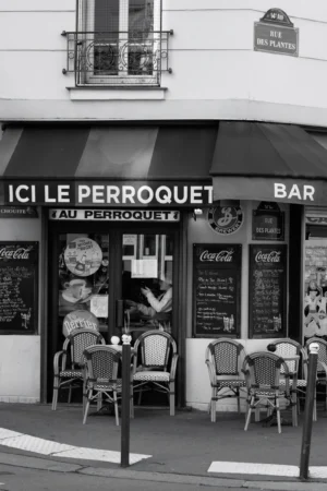 Philippe · 04/10/2025 · 2001 x 3000 Cette photographie en noir et blanc montre la façade d’un petit commerce nommé **« Ici Le Perroquet »**, avec une enseigne indiquant aussi un bar. Le sujet principal est l’entrée du café, avec ses tables et chaises en extérieur disposées devant.La composition est symétrique, avec des éléments bien alignés : tables, enseignes et menu visible. Le cadrage est serré sur la devanture, capturant une partie de la rue. La lumière est naturelle, douce, sans ombres marquées, créant une ambiance calme et rétro. Les tons gris dominent, accentuant le charme vintage du lieu. L’ambiance est conviviale et intemporelle.