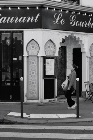 Philippe · 04/10/2025 · 2001 x 3000 Cette photographie en noir et blanc met en scène un homme marchant devant un restaurant nommé **« Le Gourbi »**. Le sujet principal est cet homme, en mouvement, avec une posture légèrement courbée.La composition est équilibrée, avec un cadrage serré sur la façade ornée de motifs géométriques et de lettres stylisées. La lumière est douce, créant des contrastes nets entre ombres et clartés. L’ambiance est urbaine et intemporelle, renforcée par le monochrome. Le style évoque un réalisme documentaire, typique des clichés de rue classiques. L’atmosphère est à la fois banale et évocatrice, mêlant quotidien et charme rétro.