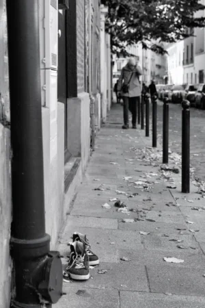 Philippe · 04/10/2025 · 2001 x 3000 Cette photographie en noir et blanc met en scène un **pied chaussé d’une basket** posé sur un trottoir. La **composition** est simple et centrée, avec une perspective légèrement basse. Le **cadrage** est serré, isolant le détail des chaussures parmi des éléments urbains (bâtiments, poubelles, feuilles).La **lumière** est naturelle, douce et diffuse, créant des contrastes modérés. Les **couleurs dominantes** sont les tons de gris, typiques du noir et blanc, renforçant l’aspect mélancolique.Le **style** est documentaire et réaliste, évoquant une **ambiance** urbaine solitaire et intime, presque poétique.