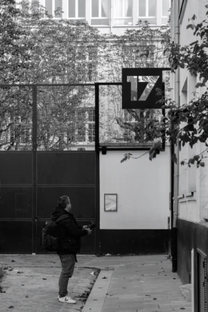 Philippe · 04/10/2025 · 2001 x 3000 Cette photographie en noir et blanc met en scène une personne marchant dans une rue urbaine. Le sujet principal est cette silhouette vue de dos, vêtue d’un manteau et portant un sac.La composition est simple et épurée, avec un cadrage serré sur la personne et un arrière-plan minimaliste : un mur, une enseigne "TZ" et une clôture. La lumière est douce, légèrement tamisée, créant des contrastes modérés. L’ambiance est calme et introspective, renforcée par le monochrome qui accentue les textures et les ombres. Le style est documentaire et sobre, évoquant une atmosphère urbaine mélancolique.