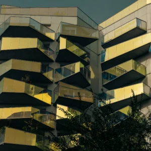 Don · 18/10/2025 · 2048 x 2048 Cette photographie met en scène un immeuble moderne aux balcons en saillie. La composition joue sur les lignes horizontales et verticales, créant un effet géométrique. Le cadrage serré accentue la structure architecturale.La lumière, douce et naturelle, semble provenir d’un angle bas, soulignant les contrastes. Les couleurs dominantes sont les tons beiges et jaunes, rehaussés par des reflets dorés. L’ambiance est à la fois minimaliste et dynamique, évoquant une architecture contemporaine et épurée. L’ombre des arbres ajoute une touche organique à la scène.