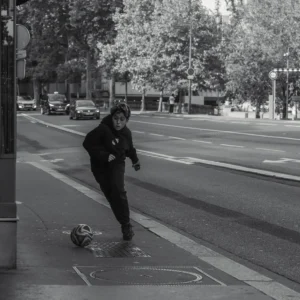 Don · 18/10/2025 · 2048 x 2048 Cette photographie en noir et blanc met en scène un jeune homme comme sujet principal, en action avec un ballon de football. La composition montre une rue urbaine avec des arbres, des voitures et des bâtiments en arrière-plan.Le cadrage est serré sur le sujet, capturant son mouvement dynamique. La lumière semble naturelle, probablement en journée, avec des contrastes marqués. Les couleurs dominantes, bien que monochromes, jouent sur les nuances de gris.Le style est documentaire et spontané, évoquant une ambiance authentique et vivante, presque sportive et insouciante.