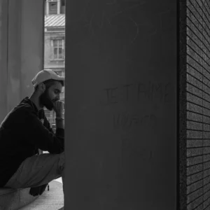 Don · 18/10/2025 · 2048 x 2048 Cette photographie met en scène un homme assis dans un recoin sombre, comme en retrait. Le sujet principal est cet homme, pensif, avec une posture légèrement courbée et les mains jointes sous son menton.La composition joue sur le cadrage serré et l’angle bas, créant une impression d’intimité et de solitude. La lumière est faible et contrastée, accentuant les ombres et donnant un côté dramatique.Les couleurs dominantes sont les tons de gris, typiques d’une photo en noir et blanc, renforçant l’ambiance mélancolique et contemplative. Le style est réaliste et sobre, évoquant une atmosphère introspective et urbaine.