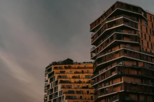 Don · 18/10/2025 · 2048 x 1365 Cette photographie met en scène deux immeubles modernes en hauteur. Le sujet principal est l’architecture contemporaine avec ses balcons et ses lignes géométriques. La composition joue sur la symétrie et la verticalité, renforcée par le cadrage serré.La lumière, douce et dorée, provient probablement du coucher de soleil, créant des ombres et des contrastes. Les couleurs dominantes sont les tons chauds (oranges, bruns) et les gris, avec des touches de bleu dans le ciel crépusculaire.Le style est épuré et urbain, évoquant une ambiance à la fois moderne et mélancolique, presque poétique.