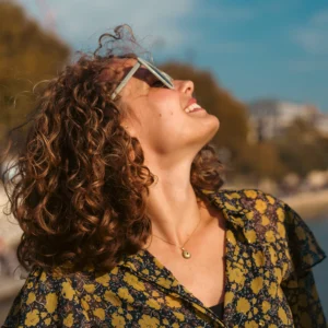 Don · 11/10/2025 · 3481 x 3481 Cette photographie met en scène une femme comme sujet principal, les yeux fermés et la tête renversée en arrière, comme savourant un moment de plaisir ou de détente. Le cadrage est serré, centré sur son visage et ses cheveux bouclés.La lumière naturelle, douce et légèrement latérale, éclaire son visage, créant des contrastes subtils. Les couleurs dominantes sont chaudes : les tons dorés et verts de son vêtement à motifs floraux, contrastant avec le bleu du ciel.Le style est naturel et spontané, évoquant une ambiance joyeuse et insouciante.