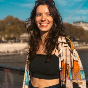 Don · 11/10/2025 · 4672 x 4672 Cette photographie met en scène une femme souriante comme sujet principal. Le cadrage est serré sur elle, avec un arrière-plan flou montrant un paysage côtier.La lumière est naturelle et douce, probablement en journée. Les couleurs dominantes sont vives : les tons chauds du vêtement coloré, contrastant avec le noir de son haut et les teintes bleutées de l’eau en arrière-plan.Le style est décontracté et estival. L’ambiance est joyeuse et détendue, évoquant une journée ensoleillée près de la mer.