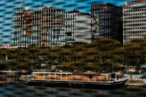 Don · 18/10/2025 · 2048 x 1365 Cette photographie met en scène un bateau ancien amarré en premier plan, partiellement caché par des arbres. L’arrière-plan révèle des bâtiments urbains, dont certains semblent en construction ou rénovation, avec des échafaudages visibles. Le cadrage est serré sur le bateau, avec une profondeur de champ réduite, créant un flou artistique.Les couleurs dominantes sont des tons froids (bleu du ciel, gris des bâtiments) contrastant avec les teintes chaudes du bateau et des arbres. La lumière semble naturelle, avec des reflets et des ombres douces, évoquant une ambiance à la fois nostalgique et moderne. Le style est photographique, presque cinématographique, avec une touche poétique.