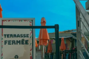 Don · 18/10/2025 · 2048 x 1365 Cette photographie montre une terrasse de café fermée, indiquée par le panneau "Terrasse Fermée". Le sujet principal est un parasol orange renversé, entourant des chaises et tables en métal. La composition est simple, avec un cadrage serré sur l’espace restreint de la terrasse. La lumière semble naturelle, mais légèrement voilée, créant une ambiance tamisée. Les couleurs dominantes sont les tons bleus du ciel, les oranges du parasol, et les gris métalliques des meubles. Le style est réaliste et documentaire, évoquant une ambiance calme et déserte, peut-être en fin de journée.