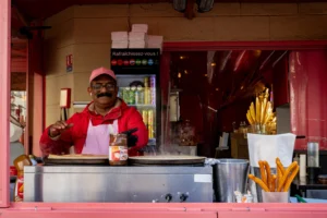 Pascal N. · 11/10/2025 · 3240 x 2160 Cette photographie montre un vendeur souriant derrière un stand de boissons et de bananes plantains. Le sujet principal est l’homme en chemise rouge, en train de boire.La composition est dynamique, avec un cadrage serré sur le vendeur et son environnement immédiat. La lumière est naturelle et légèrement contrastée, mettant en valeur les détails.Les couleurs dominantes sont les tons chauds : rouge, jaune (des bananes), et blanc. L’ambiance est chaleureuse et vivante, évoquant un marché local ou une rue animée. Le style est réaliste et spontané.