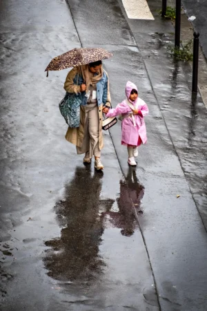 AnJo · 03/10/2025 · 1280 x 1920 Cette photographie capture une scène urbaine sous la pluie. Le **sujet principal** est une femme tenant un parapluie à motifs et une fillette en manteau rose, toutes deux marchant sur un trottoir mouillé.La **composition** est simple et naturelle, avec un **cadrage serré** mettant en valeur les deux personnages. La **lumière** est tamisée, typique d’un jour gris et pluvieux, créant des reflets sur le sol.Les **couleurs dominantes** sont les tons froids (gris, bleu) et quelques touches chaudes (rose du manteau). Le **style** est documentaire et réaliste, évoquant une **ambiance** intimiste et mélancolique, soulignant la douceur et la vulnérabilité face à la pluie.