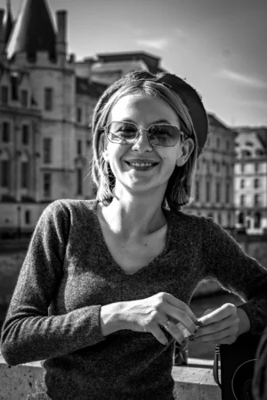 AnJo · 11/10/2025 · 1280 x 1920 Cette photographie en noir et blanc met en scène une jeune femme comme sujet principal, souriante et détendue. Elle porte un pull épais et des lunettes, tenant un objet à la main.La composition est simple et centrée, avec un cadrage serré sur son visage et sa poitrine. L’arrière-plan flou montre des bâtiments, suggérant un cadre urbain.La lumière est douce et naturelle, créant des contrastes modérés. Le style est documentaire et spontané, évoquant une ambiance chaleureuse et authentique, renforcée par les tons de gris et les ombres délicates.