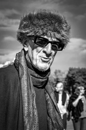 AnJo · 11/10/2025 · 1280 x 1920 Cette photographie met en scène un homme âgé comme sujet principal, vêtu d’un manteau, d’un foulard et d’un chapeau à fourrure, avec des lunettes de soleil. Le cadrage est serré, centré sur son visage, isolant le sujet du fond flou et légèrement estompé.La lumière semble naturelle, contrastée, créant des ombres marquées. En noir et blanc, les tons varient du clair au foncé, accentuant les textures et les détails. Le style est documentaire et intime, évoquant une ambiance à la fois sobre et expressive, avec une touche de mélancolie et de caractère.