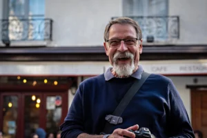 AnJo · 11/10/2025 · 1920 x 1280 Cette photographie met en scène un homme âgé comme sujet principal, souriant et portant des lunettes, un pull bleu et un appareil photo autour du cou. Le cadrage est serré, centré sur lui, avec un arrière-plan flou montrant une façade de bâtiment et des enseignes.La lumière est naturelle, douce et légèrement contrastée, créant une ambiance chaleureuse. Les couleurs dominantes sont les tons bleus du pull et les teintes neutres de la peau et des bâtiments. Le style est documentaire et spontané, évoquant une scène de rue authentique et décontractée. L’ambiance est conviviale et humaine.