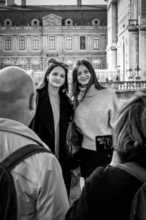 AnJo · 11/10/2025 · 1280 x 1920 Cette photographie en noir et blanc met en scène deux femmes souriantes comme sujets principaux, posant devant un bâtiment classique. La composition est symétrique, avec les deux femmes au centre. Le cadrage est serré sur elles, tout en incluant partiellement d'autres personnes en arrière-plan.La lumière est douce et naturelle, mettant en valeur les visages. Les tons dominants sont les gris et noirs, typiques du noir et blanc, avec quelques contrastes marqués. Le style est documentaire et spontané, capturant un moment authentique. L’ambiance est chaleureuse et conviviale, évoquant une visite touristique ou un moment de partage.