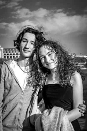 AnJo · 11/10/2025 · 1280 x 1920 Cette photographie en noir et blanc met en scène deux jeunes posant côte à côte. Le sujet principal est ce couple souriant, avec une composition serrée et équilibrée. Le cadrage est portrait, centré sur leurs visages et épaules.La lumière douce, probablement naturelle, adoucit leurs traits et crée des contrastes subtils. L’ambiance est chaleureuse et intime, renforcée par leur proximité et leurs expressions détendues. Le style est sobre et élégant, typique d’un portrait artistique classique. Les couleurs, absentes en raison du noir et blanc, laissent place à des nuances de gris et de blancs.