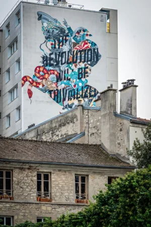 AnJo · 18/10/2025 · 1280 x 1920 Cette photographie montre un **mural** sur un immeuble, sujet principal. La composition met en scène un personnage stylisé, entouré de drapeaux et de symboles révolutionnaires, avec le texte *"Révolution will be civilized"*.Le cadrage est serré sur le bas de l’immeuble, intégrant partiellement un toit et des arbres. La lumière est naturelle, diffuse, sans ombre marquée. Les couleurs dominantes sont les **bleus, rouges et blancs**, dynamiques et contrastées.Le style est **urbain et artistique**, mêlant street art et message politique. L’ambiance est à la fois **révoltée et poétique**, évoquant un appel à une révolution pacifique.