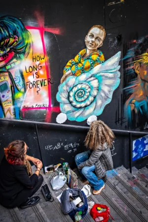 AnJo · 18/10/2025 · 1280 x 1920 Cette photographie montre des artistes urbains en train de peindre une fresque murale. Le sujet principal est le mur coloré, avec des motifs psychédéliques et des personnages stylisés. La composition inclut deux personnes assises sur des escaliers, entourées de leurs outils.Le cadrage est serré sur les artistes et la fresque en cours. La lumière est naturelle et semble provenir d’une source latérale, créant des contrastes. Les couleurs dominantes sont vives : bleus, roses, jaunes et verts.Le style est street art, dynamique et expressif, évoquant une ambiance créative et énergique.